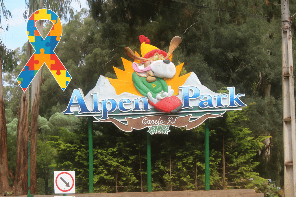 ChatGPT Image 22 de jun. de 2025, 17_03_23 Placa de entrada do Alpen Park em Canela, Rio Grande do Sul, com o mascote do parque e o laço colorido de conscientização sobre o autismo à esquerda.