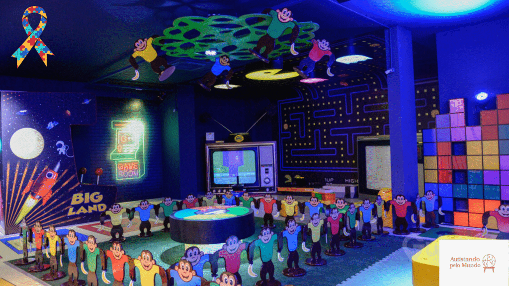 Big Land Autistic Interior do Big Land Parque Temático, com duas pessoas jogando em um fliperama gigante no estilo arcade, ao lado de um laço colorido de conscientização sobre o autismo