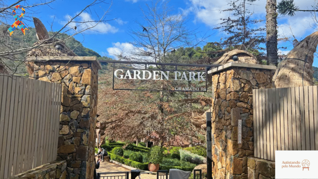 Garden Park Autistic Entrada do Garden Park em Gramado, Rio Grande do Sul, com portão de pedra e jardim verde ao fundo. No canto superior esquerdo, laço colorido do autismo com estampa de peças de quebra-cabeça simbolizando inclusão.