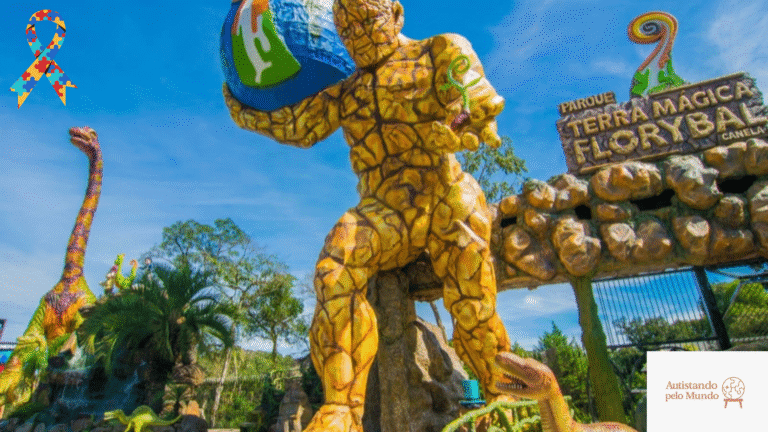Entrada do parque Terra Mágica Florybal, com estátua gigante segurando um globo e dinossauros ao redor, sob céu azul. Logo do Autistando pelo Mundo no canto inferior direito.
