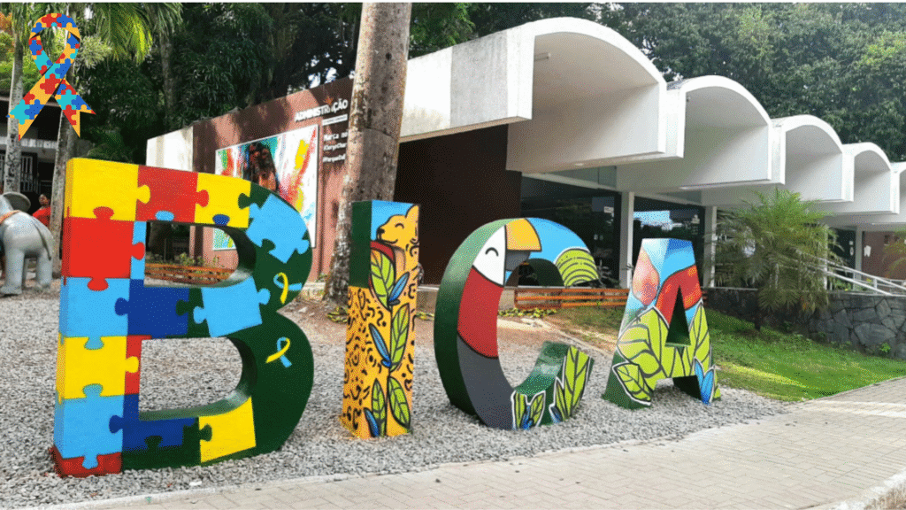 Untitled design (2) Entrada do Parque Zoobotânico Arruda Câmara em João Pessoa, com letras coloridas formando a palavra BICA, decoradas com ilustrações de animais e peças de quebra-cabeça representando inclusão.