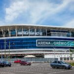 Vista externa da Arena do Grêmio em Porto Alegre, destacando a fachada azul e branca do estádio, estacionamento em primeiro plano e céu parcialmente nublado.”